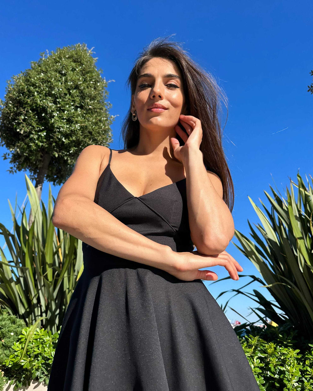 Rochie neagra elegantă scurtă cu bretele, fustă evazată și decolteu, fundal natural.